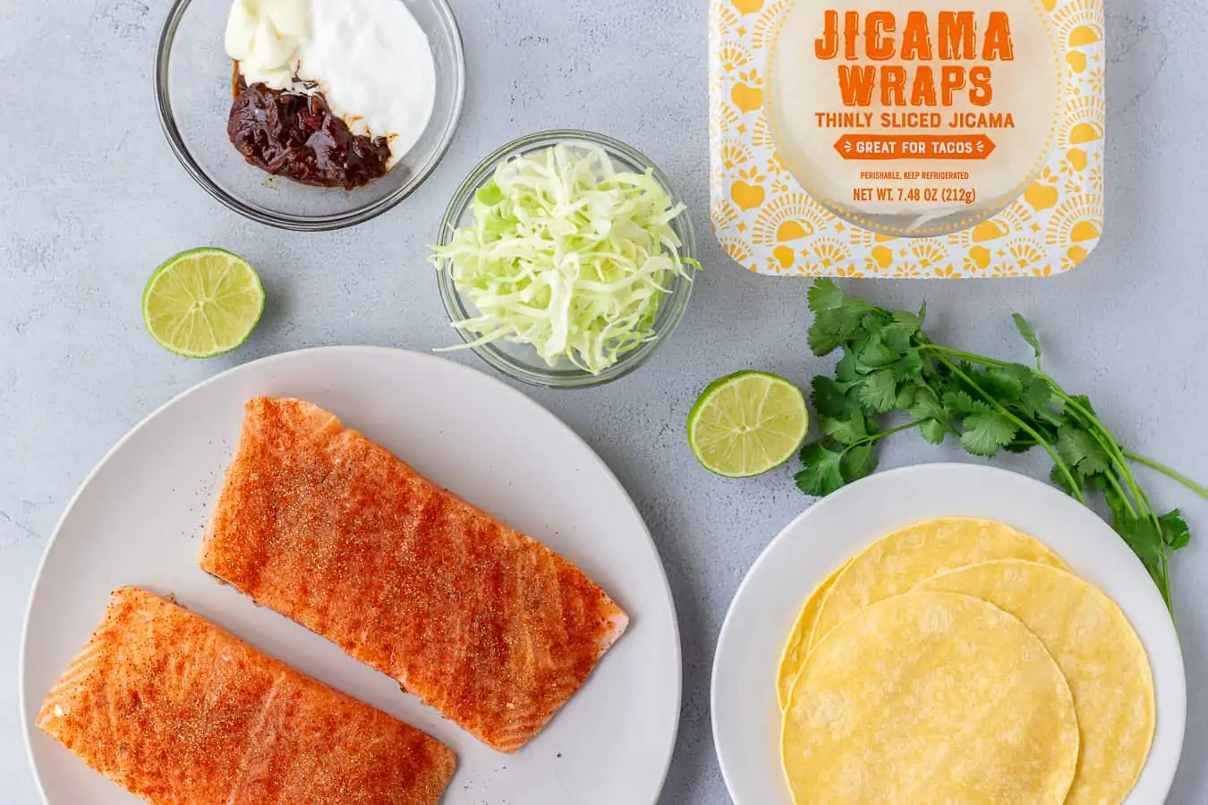 Overhead shot of ingredients, salmon fillets, shredded cabbage, corn tortillas, jicama wraps, and chipotle sauce.