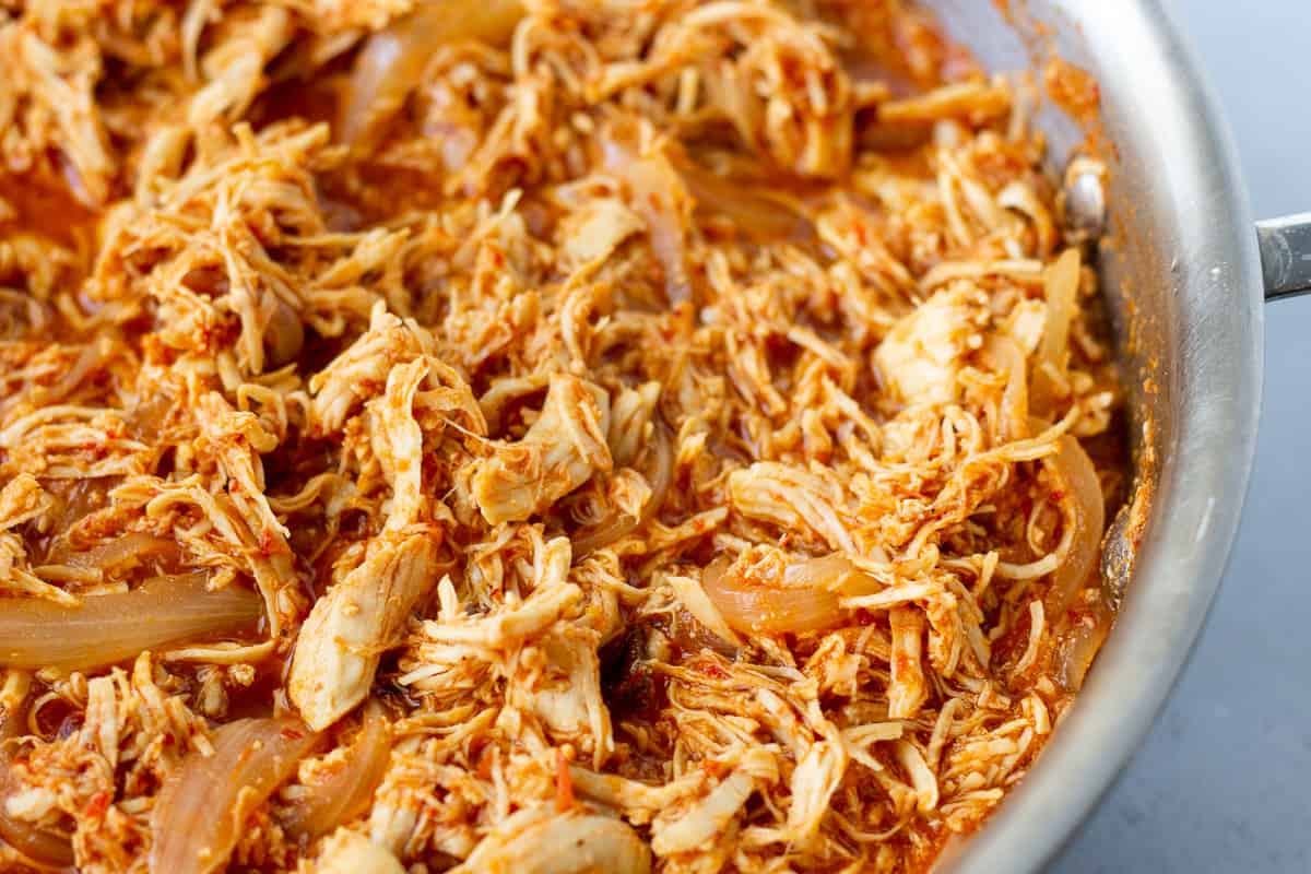 Cooked shredded chicken, onions, and red sauce in a skillet.