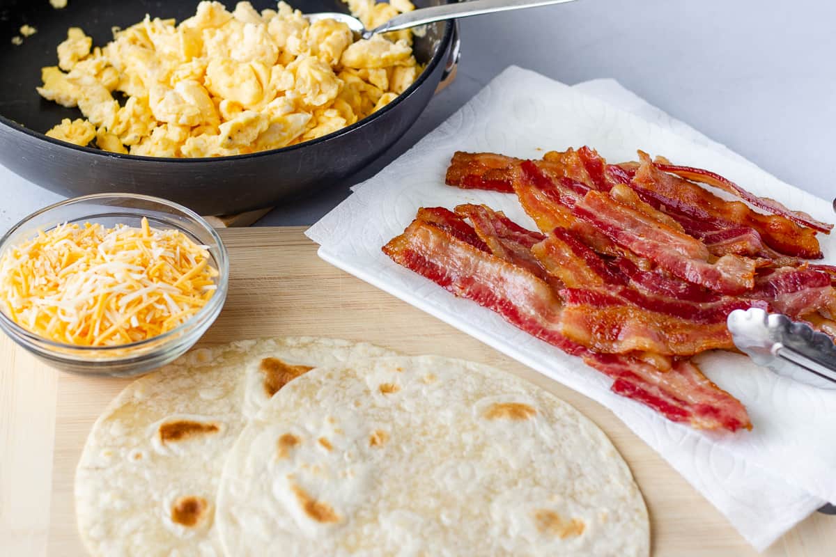 Scrambled eggs in a skillet, with slices of bacon, tortillas, and shredded cheese in a bowl.