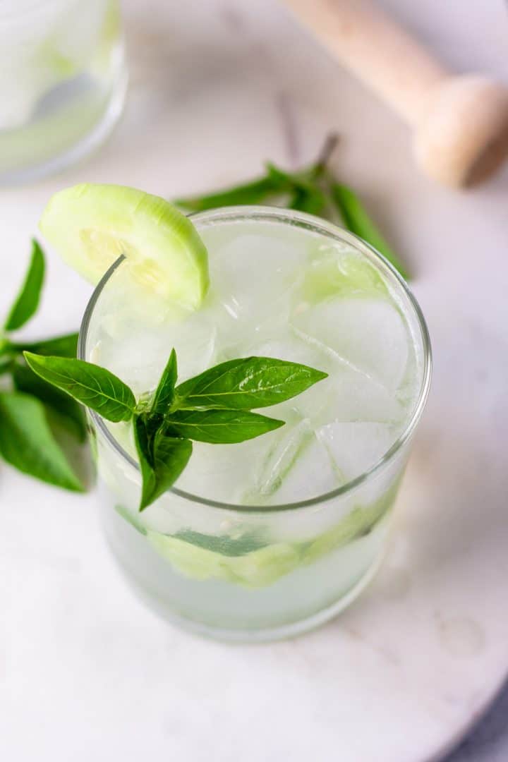 Overhead view of cocktail with basil and cucumber garnish and a muddling stick in the background.