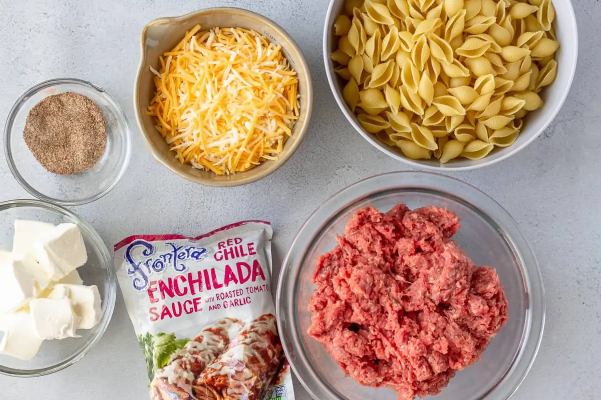 Ingredients in seperate bowls, pasta, ground beef, shredded cheese, cream cheese, and enchilada sauce.