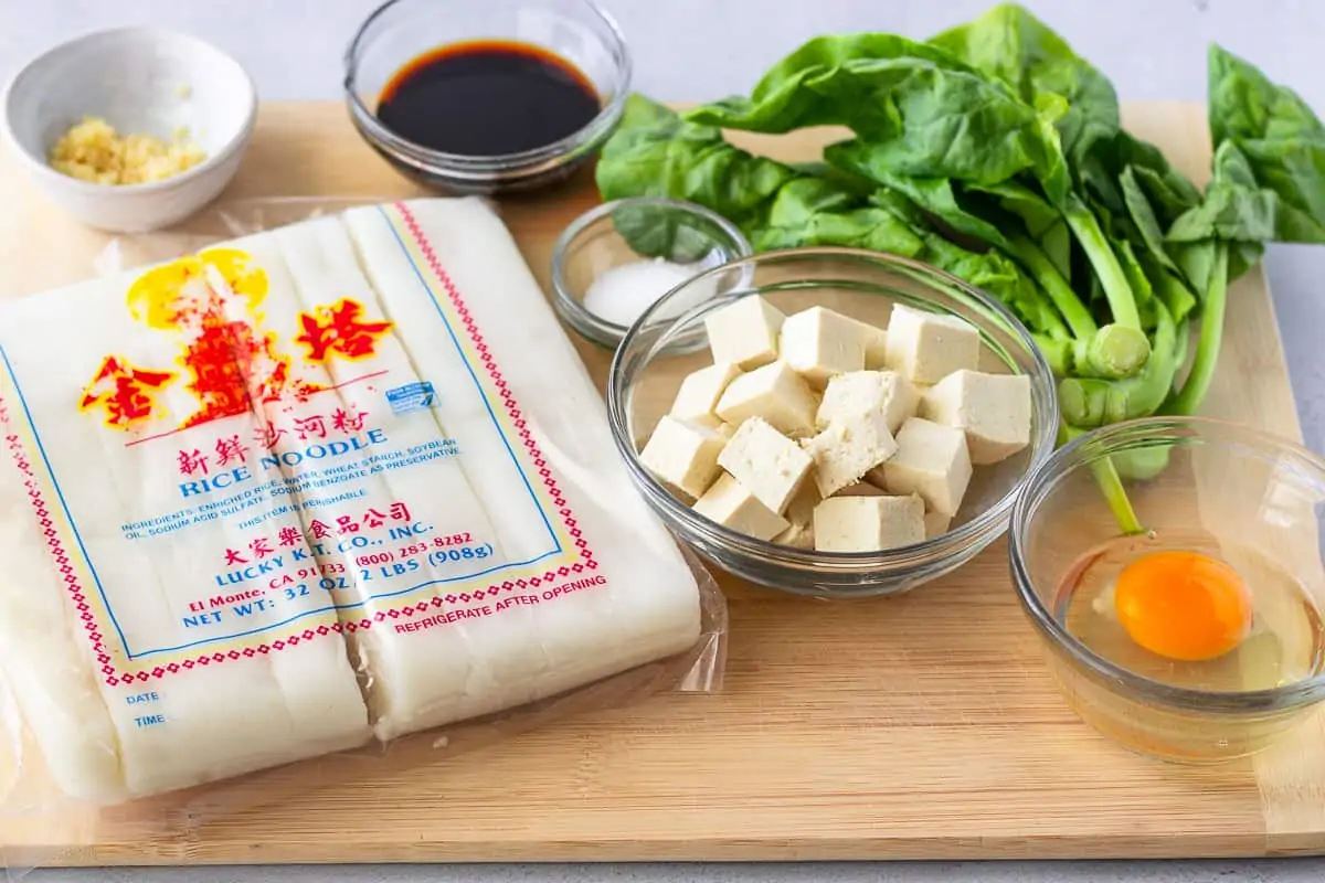 Ingredients on a cutting board, noodles, tofu, Chinese broccoli, soy sauce, and an egg.