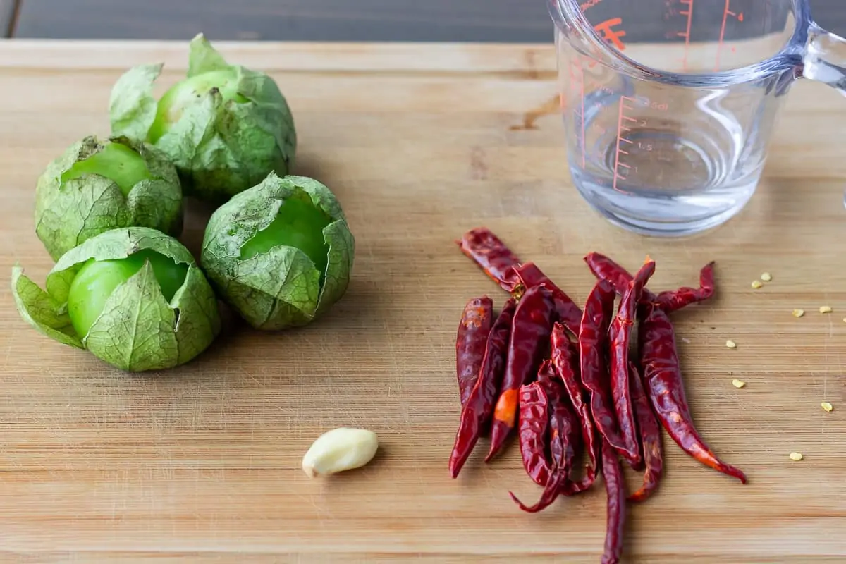 Cutting board with tomatillos, red peppers, garlic, and a glass of water.
