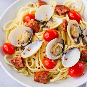 Pasta in a white bowl topped with tomatoes, clams, and sasage.