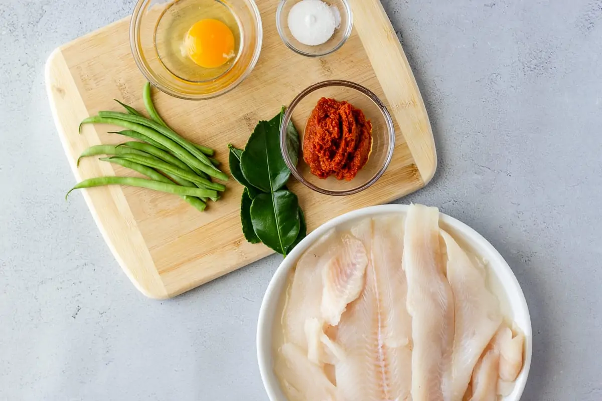 Tilapia on a plate, green beans, lime leaves, egg, curry paste, and sugar on a cutting board.