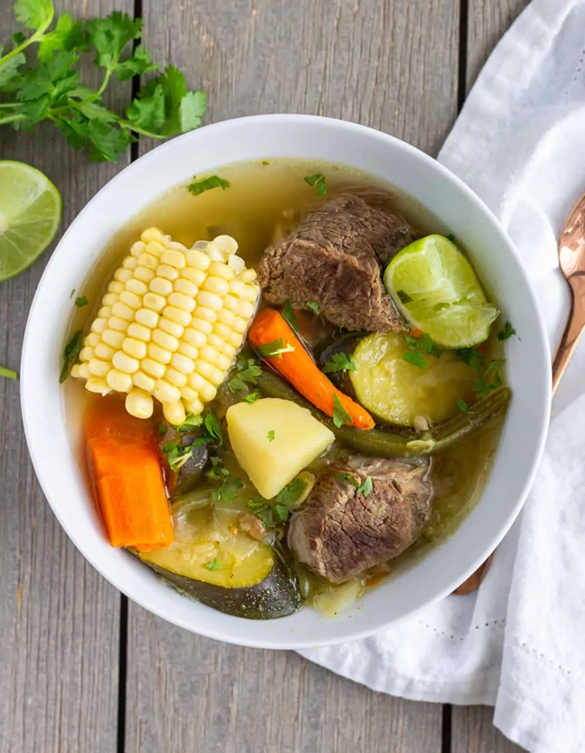 Overhead view of caldo de res in a white bowl with cilantro and lime on the side.