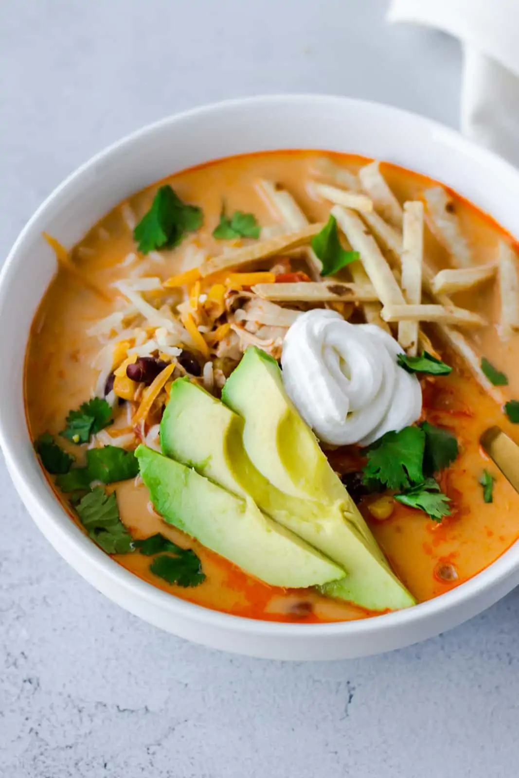 Soup in a white bowl dressed with avocado slices, sour cream, tortilla strips, and cilantro.