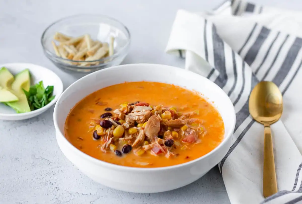 Creamy chicken tortilla soup in a white bowl with toppings in bowls on the side.