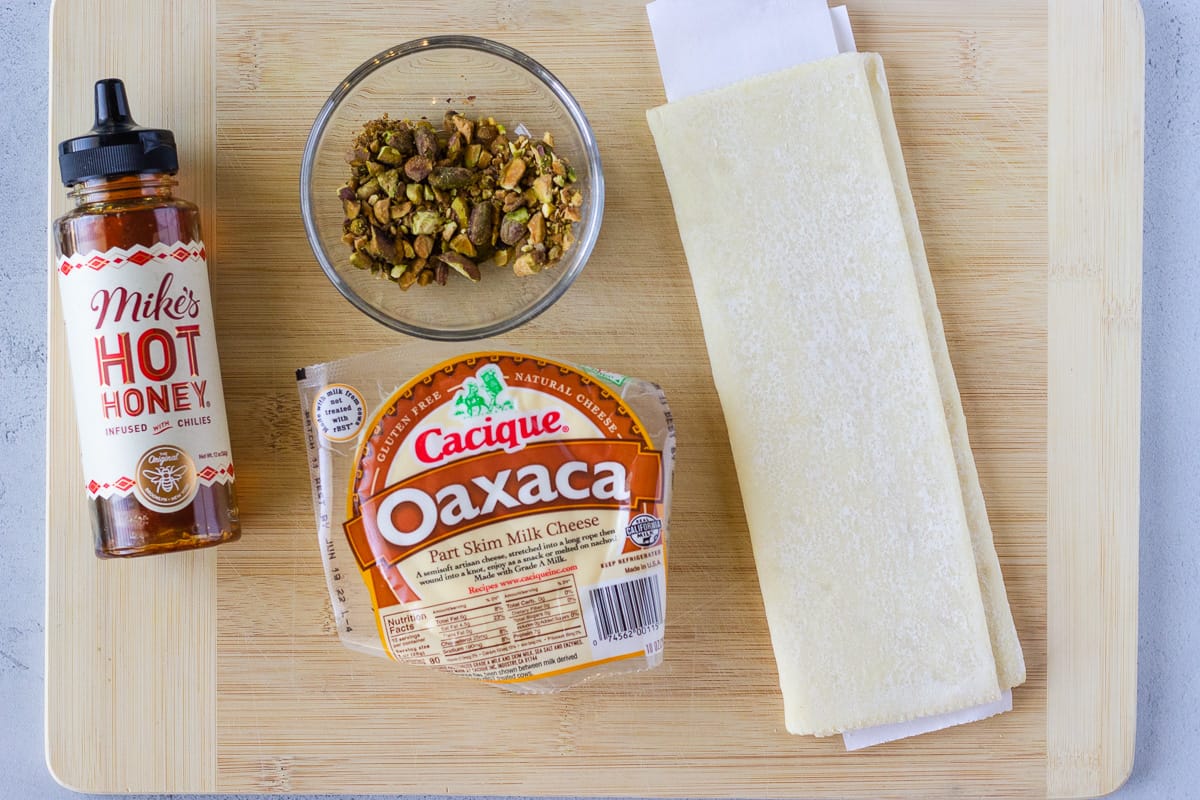 Bottle of hone honey, oaxaca cheese, crushed pistachios, and a sheet of puff pastry on a cutting board.