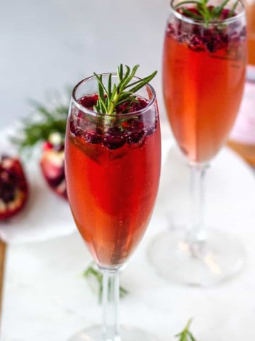 2 Champagne glasses garnished with pomegranate seeds and rosemary.