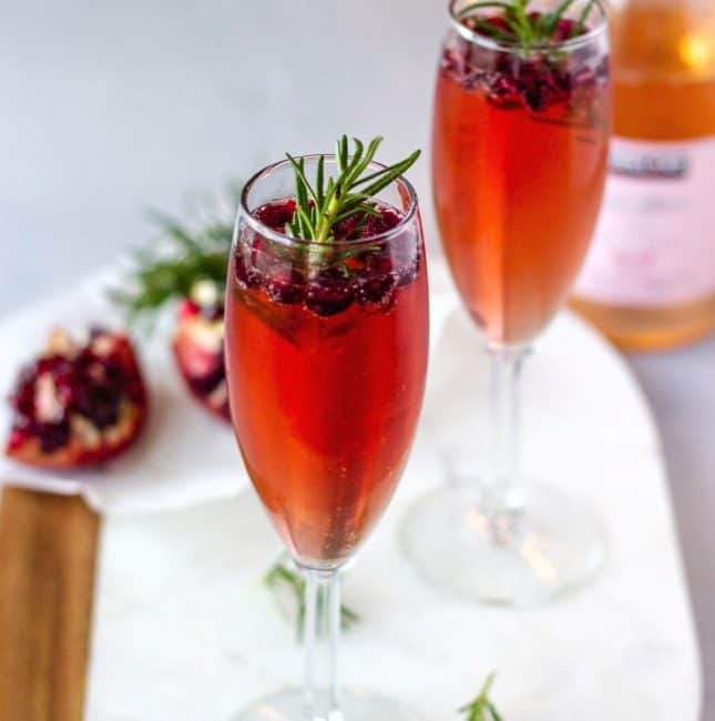 2 Champagne glasses garnished with pomegranate seeds and rosemary.