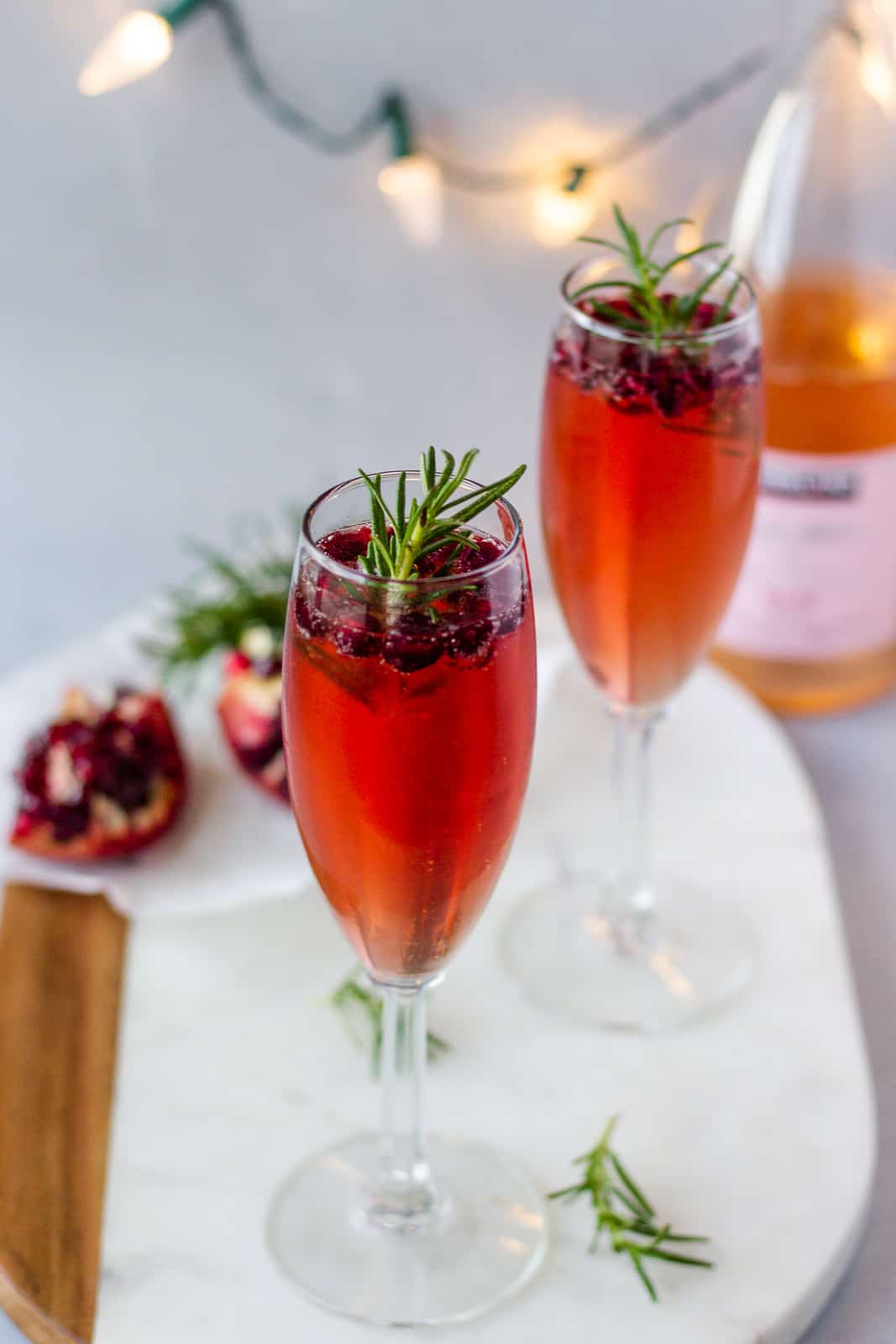 2 Champagne glasses on a board with a sliced pomegranate and twinkle lights in the background.