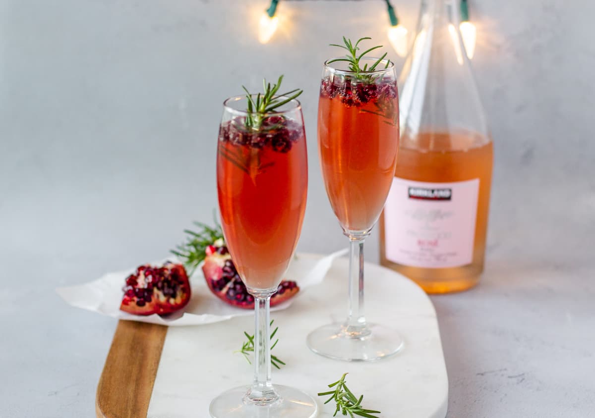 2 Champagne glasses on a board with a sliced pomegranate and bottle of prosecco in the background.