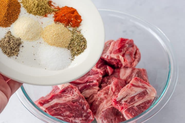 Spices being added to raw beef in a glass bowl.