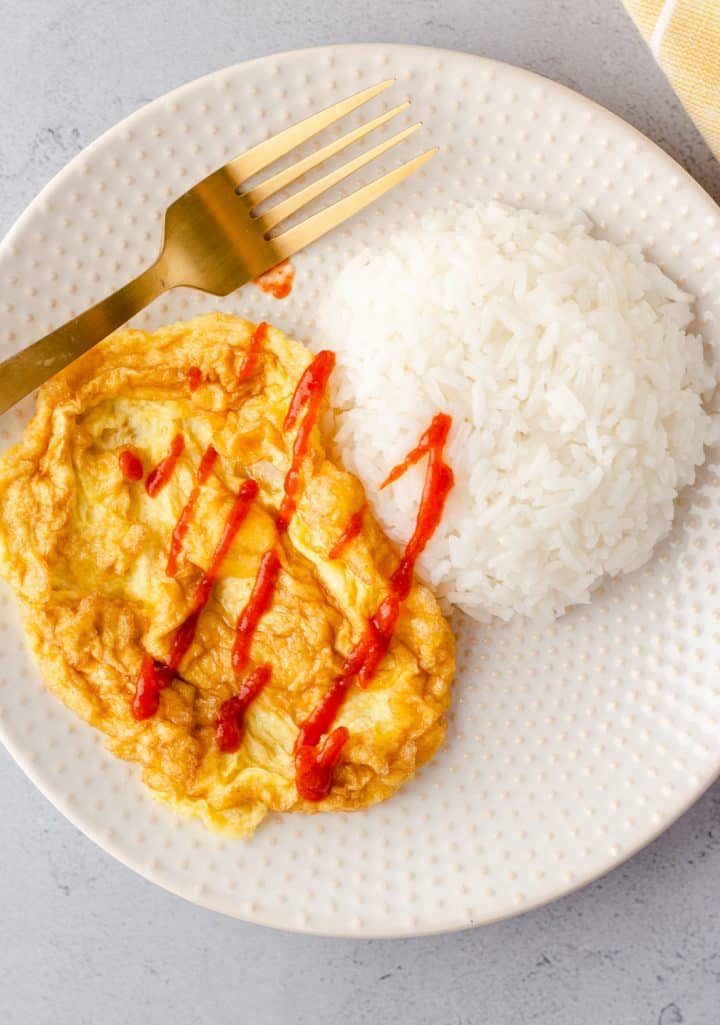 Overhead view of Thai Omelet on a plate with white rice and drizzled with siracha sauce.