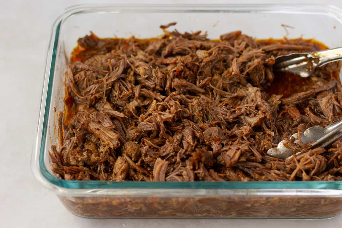 Shredded beef in a glass dish with tongs.