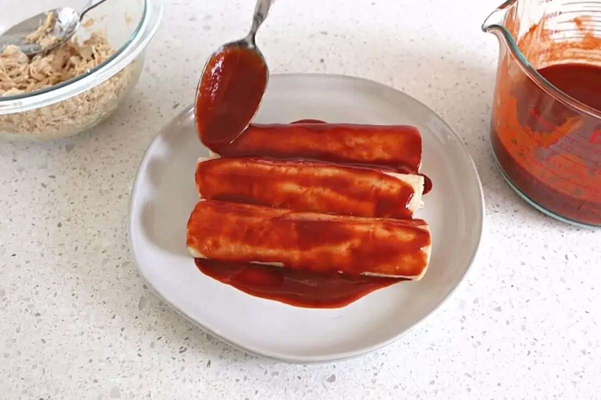 Spooning red sauce onto rolled tortillas.