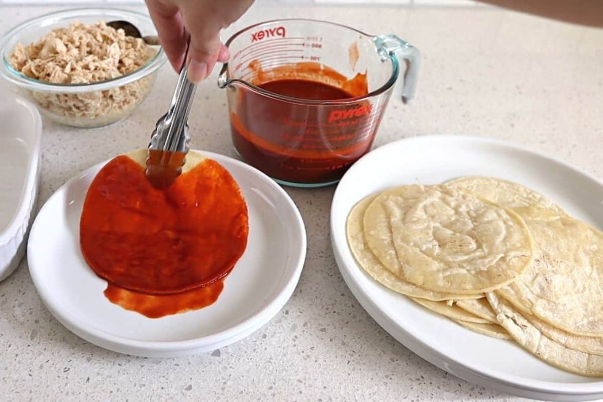 Tongs holding a tortilla dipped in red enchilada sauce.