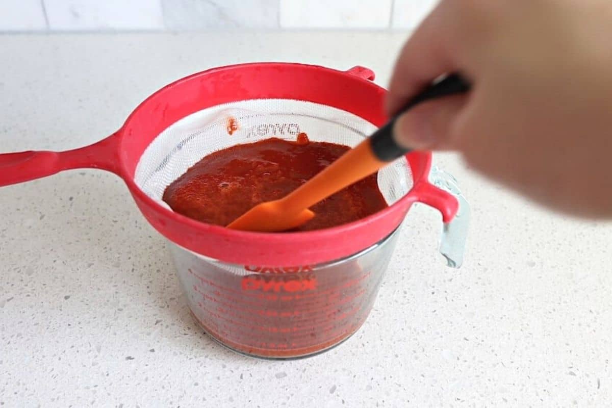 Hand holding a spatula and straining the enchilada sauce.