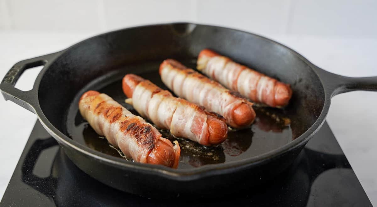 Hot dogs wrapped in bacon in a cast iron skillet.