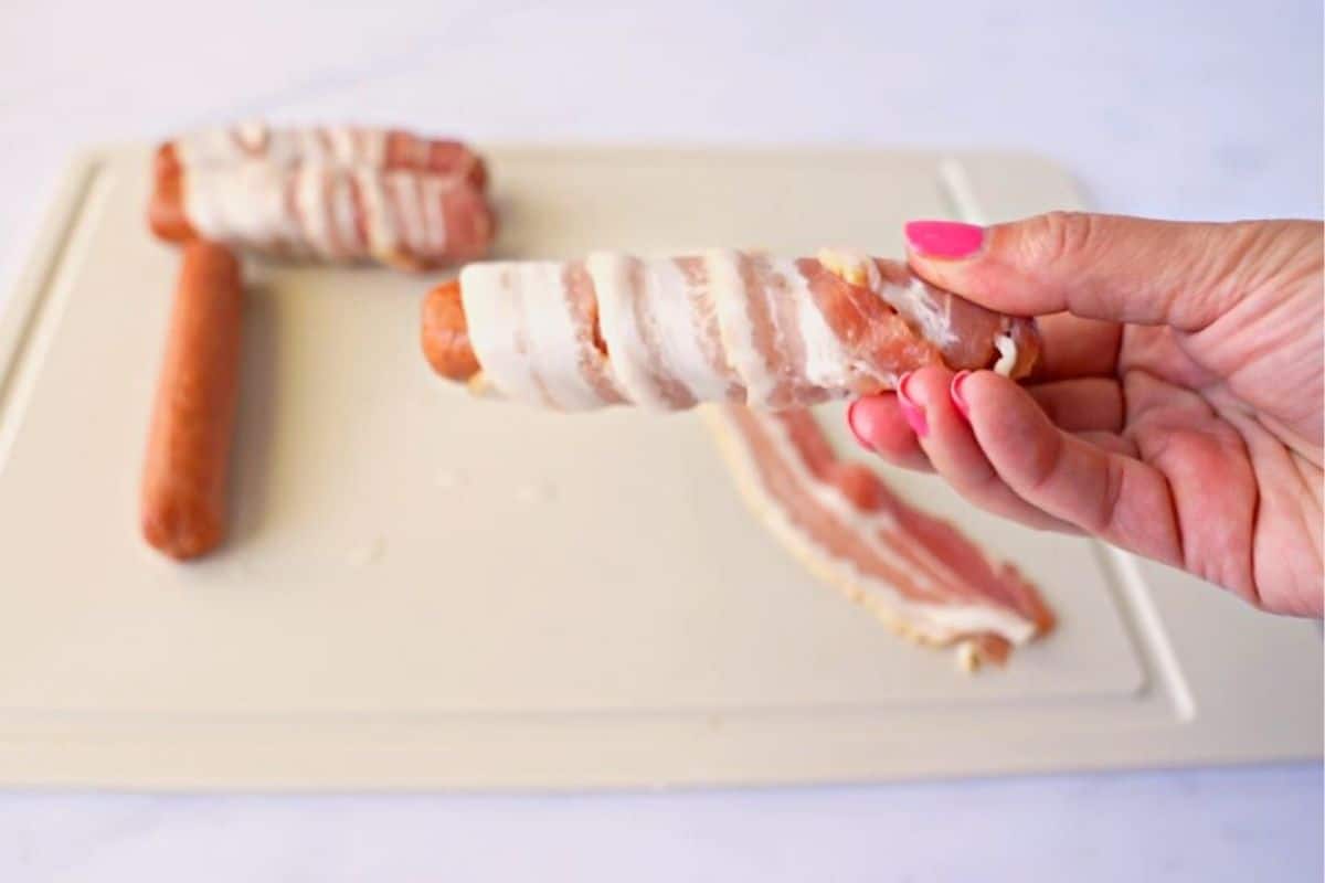 Hand wrapping a piece of bacon around a hot dog.