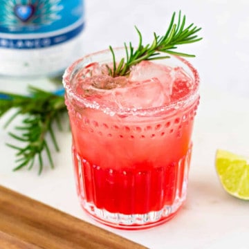 Red drink in a glass with a rosemary garnish.