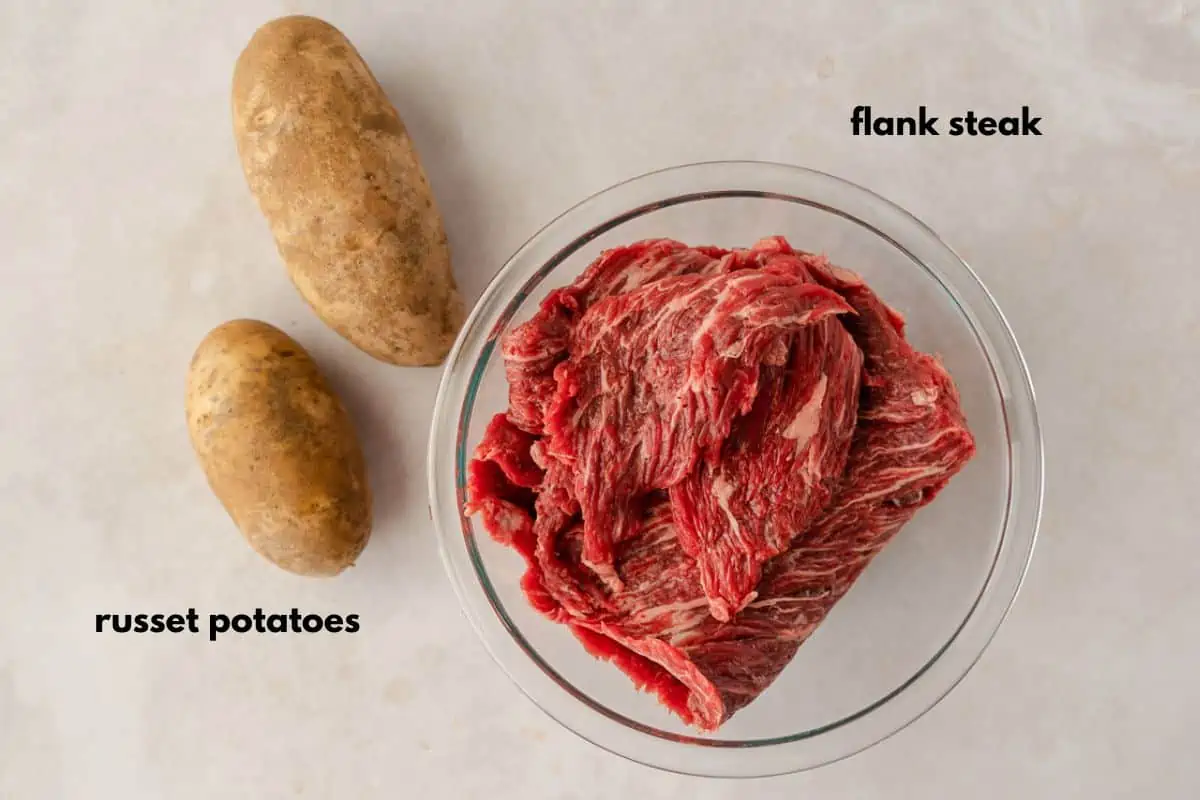 Flank steak in a bowl and 2 russet potatoes.