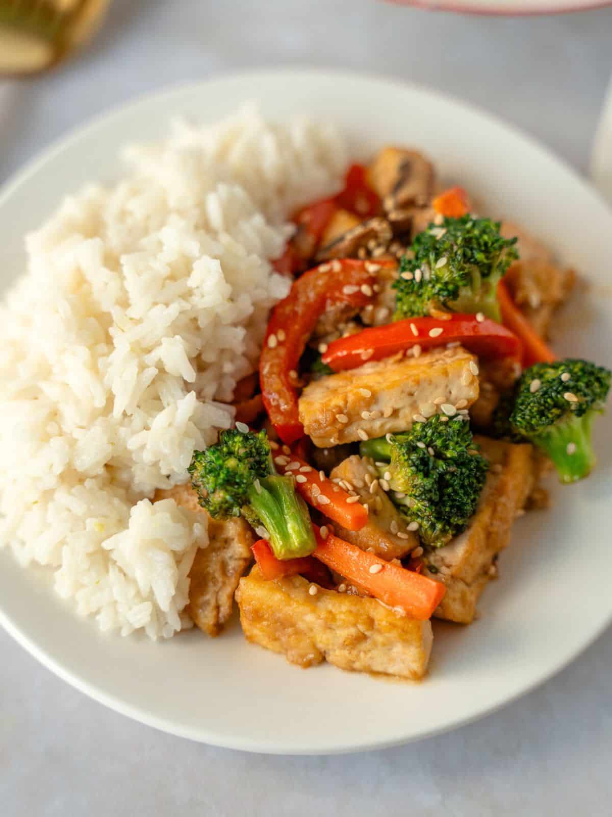 Tofu with vegetables on a plate with a side of rice.
