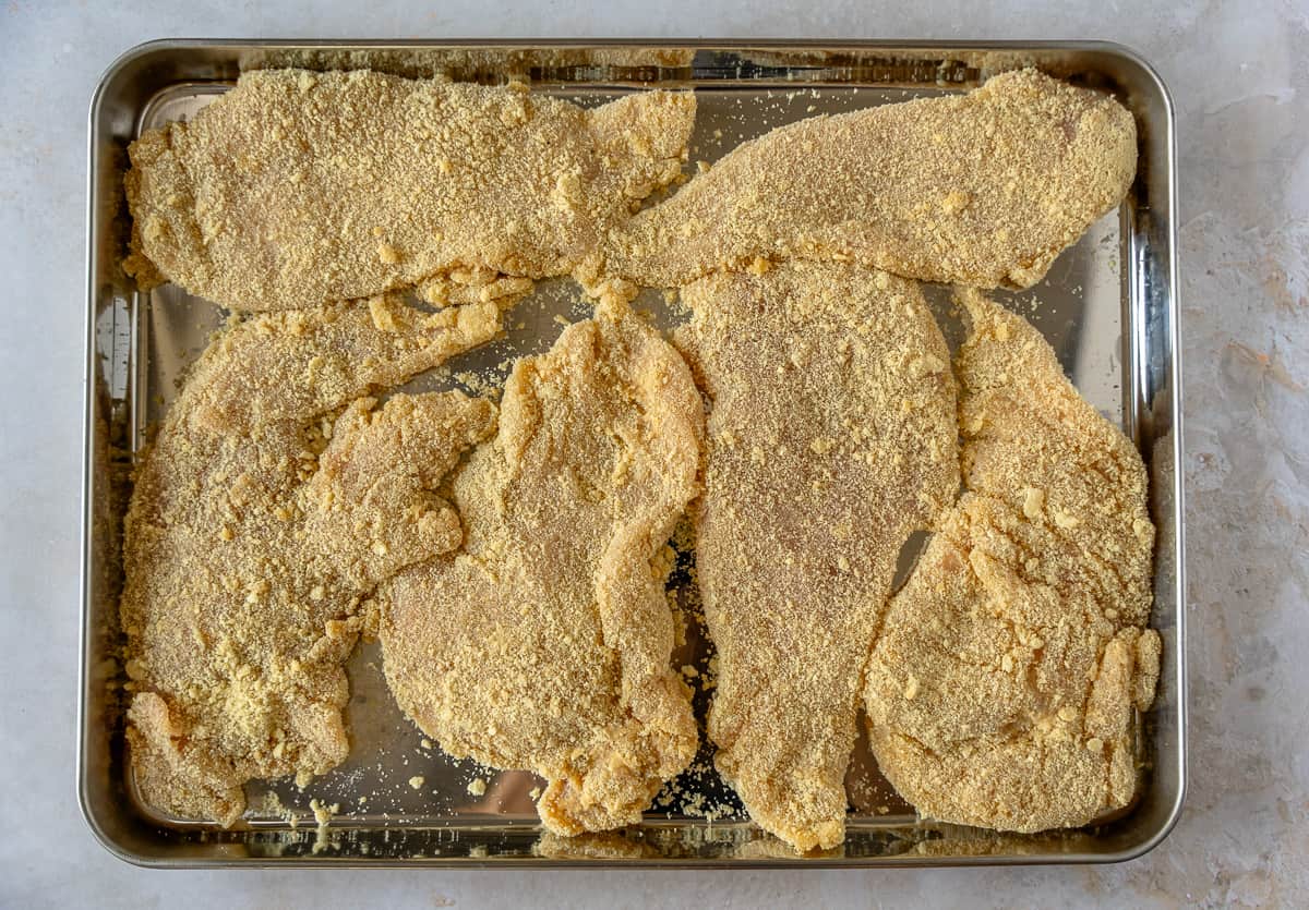 Raw chicken cutlets covered in breadcrumbs on a large baking sheet.