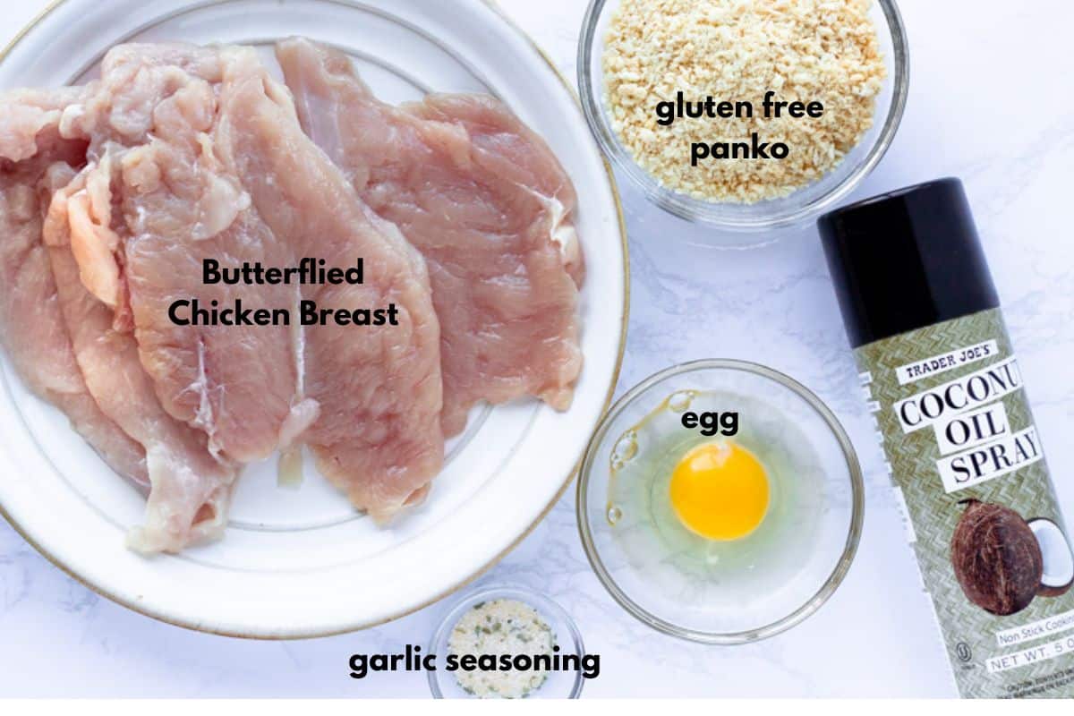 Ingredients for baked chicken milanesa labled with text. Butterflied chicken breast, gf panko, egg, garlic seasoning, and oil.