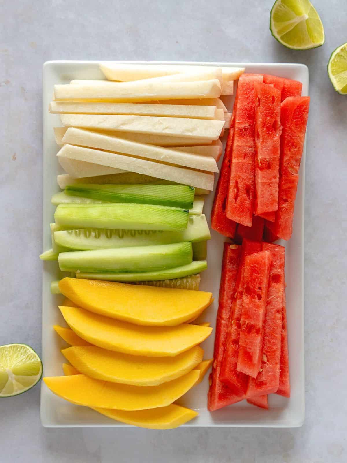 Sliced jicama, cucumber, mango, and watermelon on a rectangular plate with lime wedges on the side.