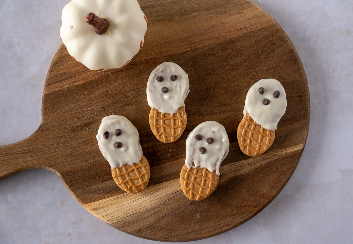 Four Nutter Butter Ghosts on a wooden board with a white pumpkin.