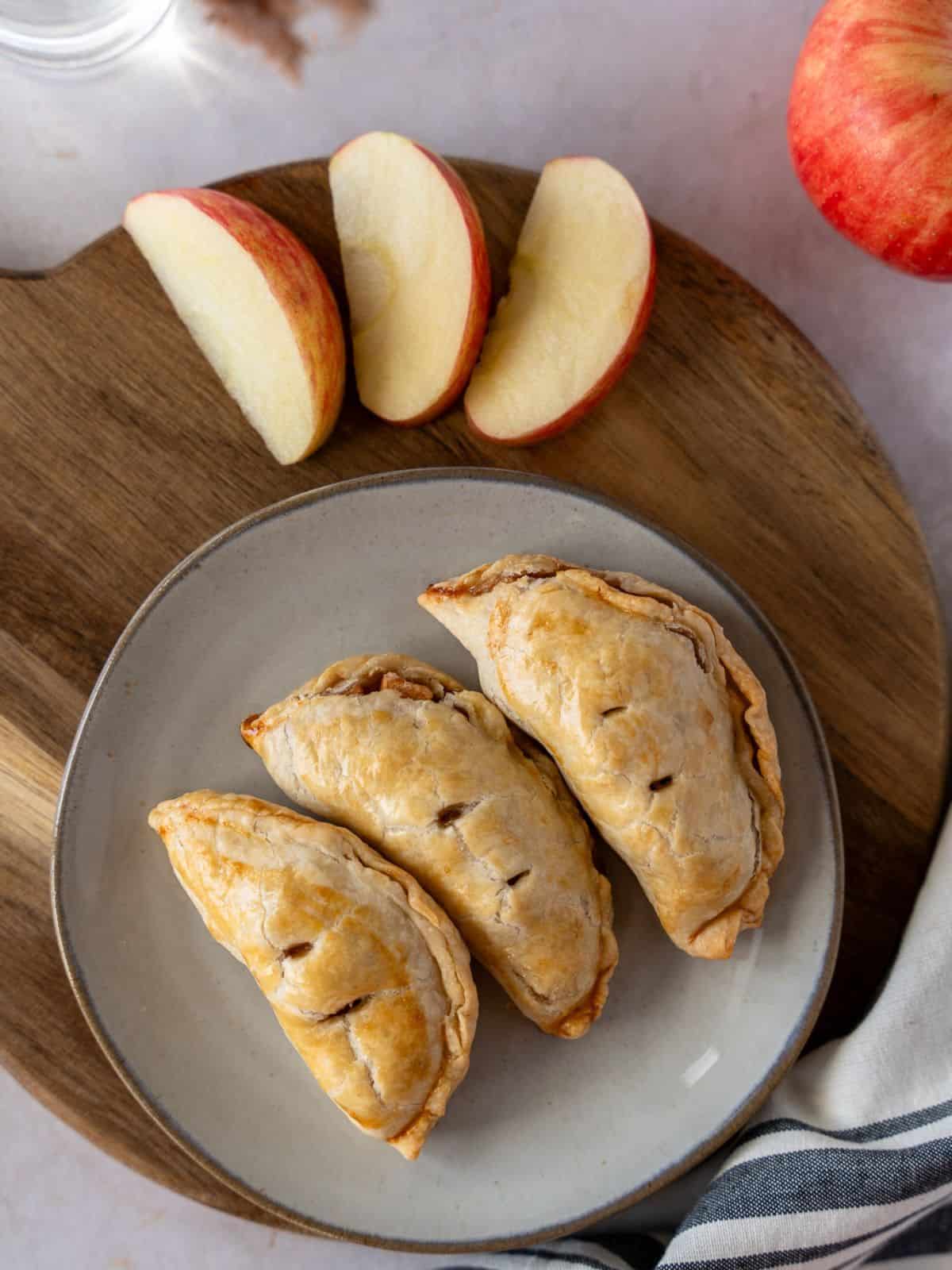 Three baked apple empanadas on a plate with some sliced apples on the side.