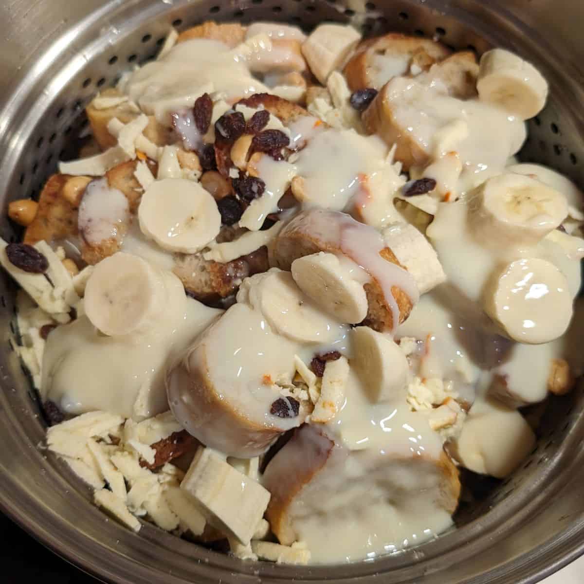 Uncooked capirotada in a pot, topped with sweetened condensed milk, banana slices, raisins, and peanuts.