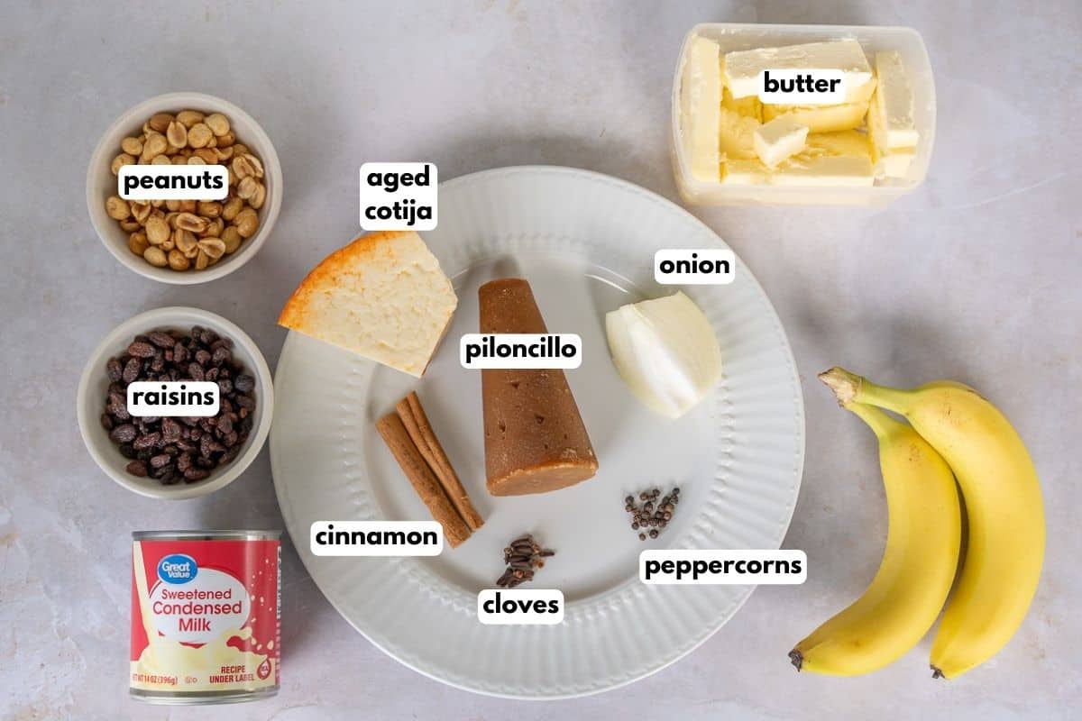 Ingredients with text, peanuts, raisins, sweetened condensed milk, aged cotija, piloncillo, cinnamon, cloves, peppercorns, onion, butter, and bananas.