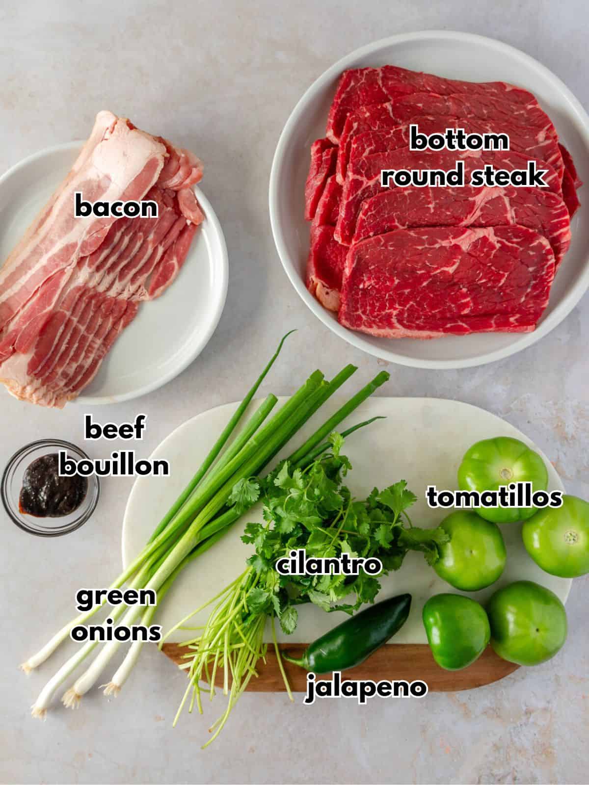 Ingredients with text, bacon, bottom round steak, beef bouillon, green onions, cilantro, jalapeno, and tamtillos.