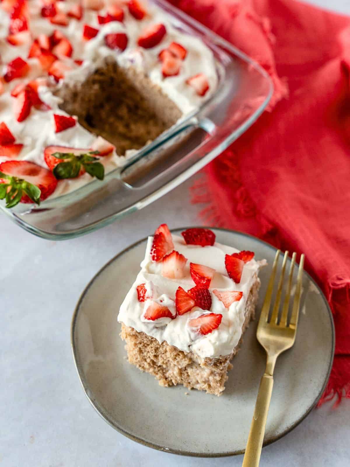 A slice of strawberry tres leches cake on a plate with the rest of the cake in the background.