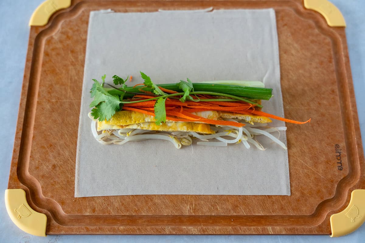 Spring roll wrapper on a cutting board with bean sprouts, eggs, carrots, green onion, and cilantro.