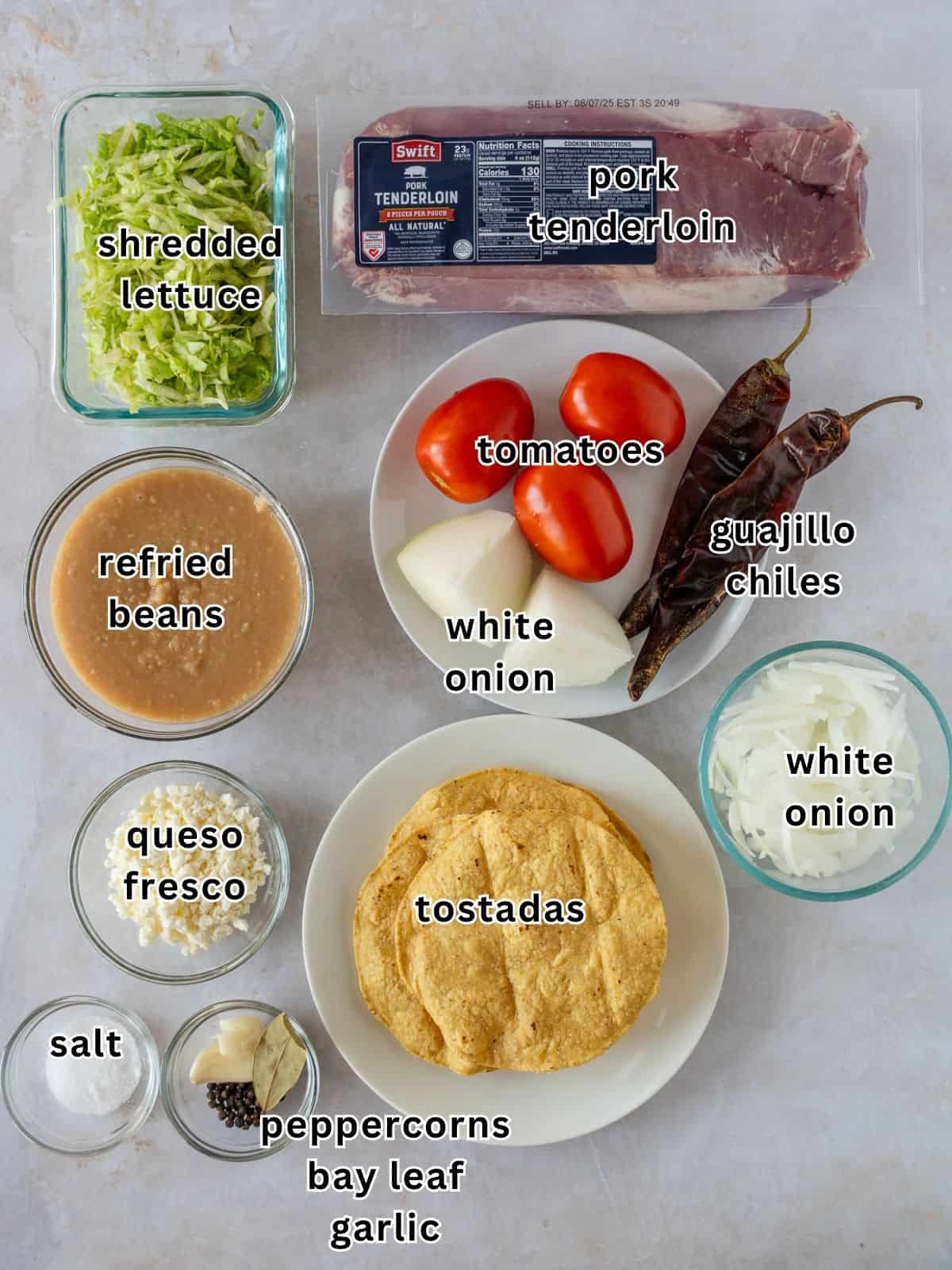 Ingredients with text- shredded lettuce, pork tenderloin, tomatoes, white onion, guajillo chiles, refried beans, queso fresco, tostadas, salt, peppercorns, bay leaf, and garlic.