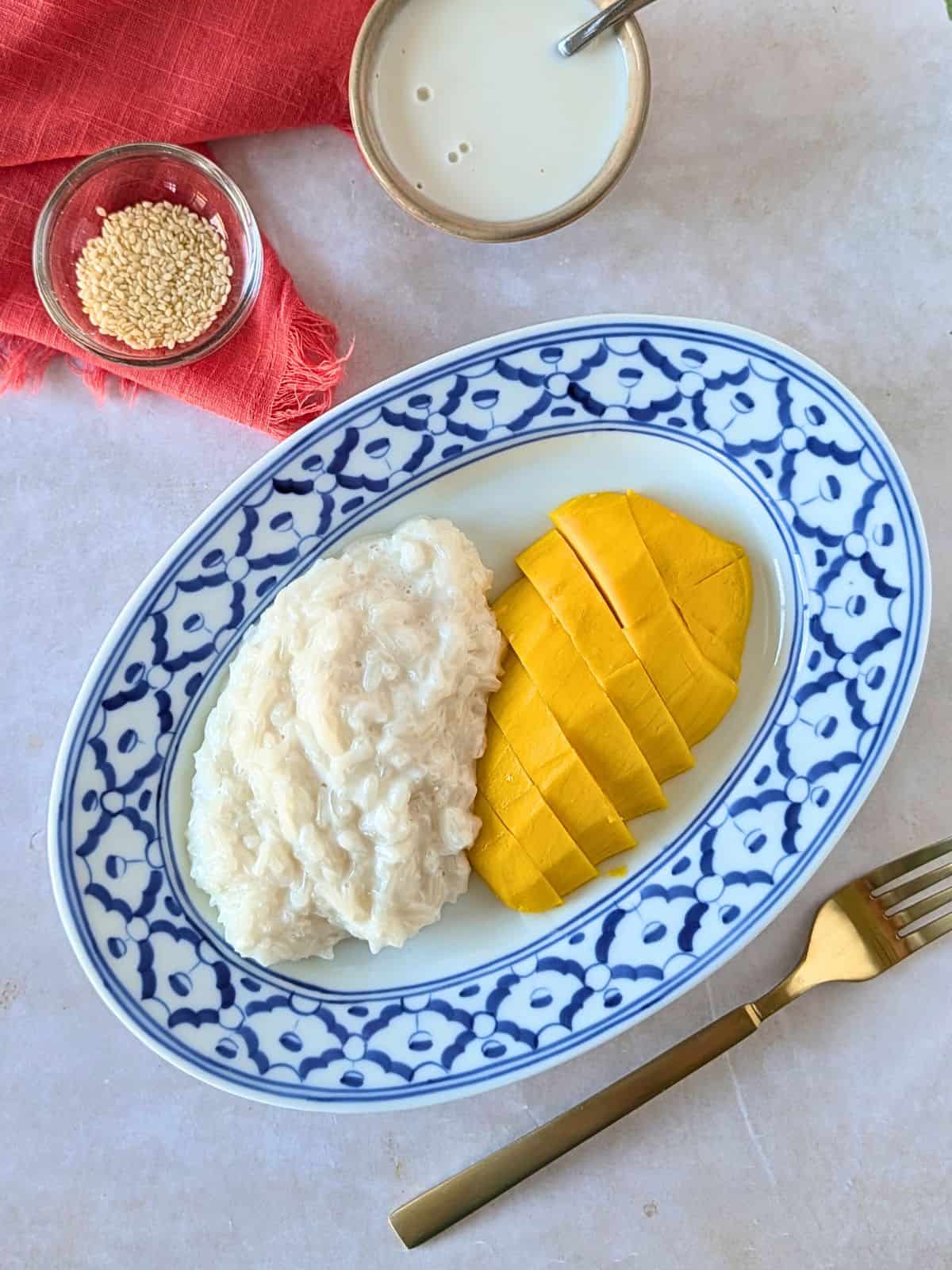 Overhead view of mango and sticky rice on a blue plate with a side of coconut milk and sesame seeds.