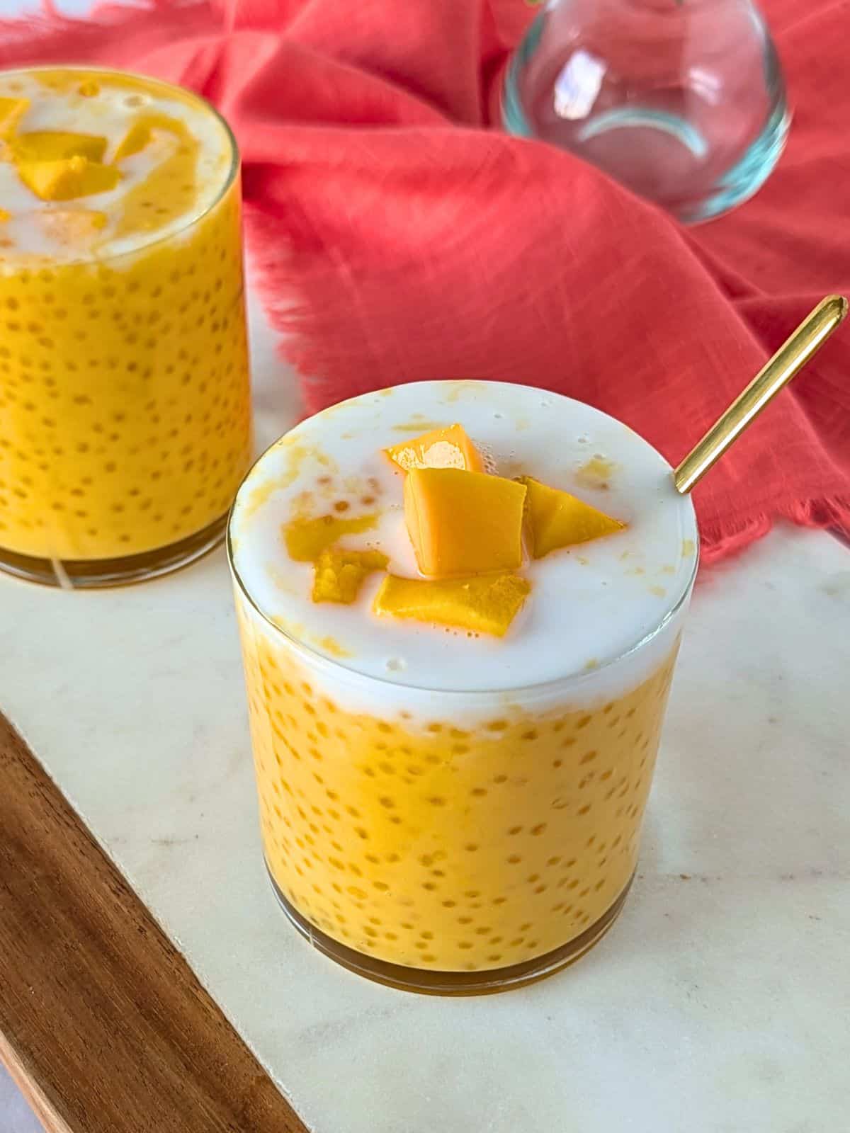 Mango sago in 2 glass cups topped with coconut milk and mango pieces.