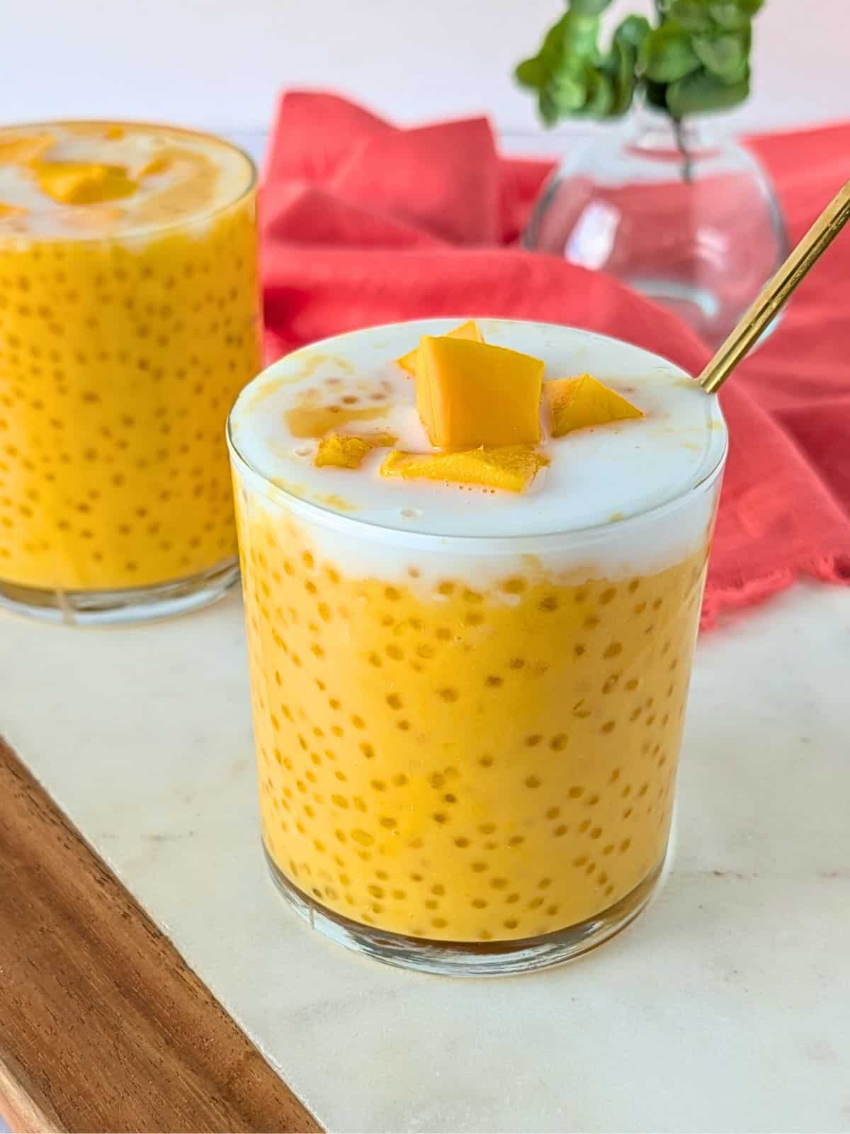 Mango sago in 2 glass cups topped with coconut milk and mango pieces.