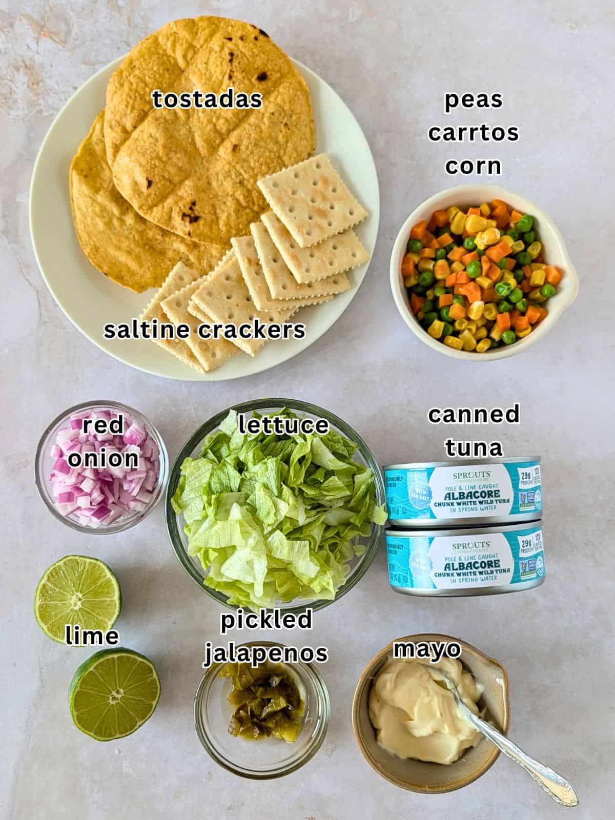 Ingredients with text- tostadas, saltine crackers, red onion, lettuce, peas, carrots, corn, tuna, lime, pickled jalapenos, and mayo.